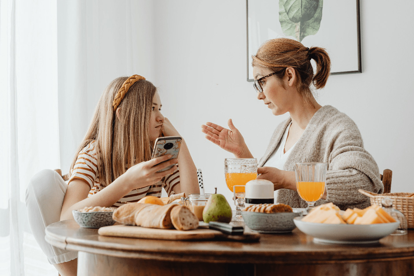 A mother gestures with her hand as her teenage daughter uses her phone on a round table adorned with bread, cheese, fruit, and juices