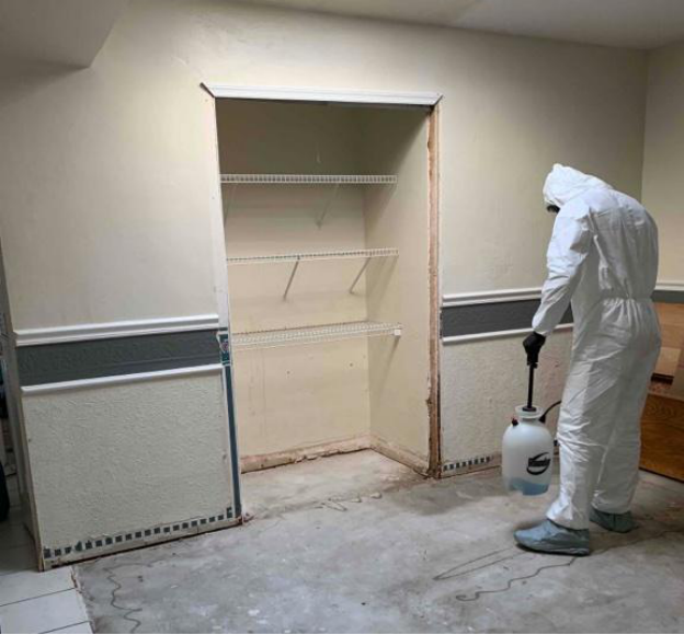 A water damage restoration specialist stands in protective gear in front of an empty shelf in an empty room, holding a pressure spray bottle