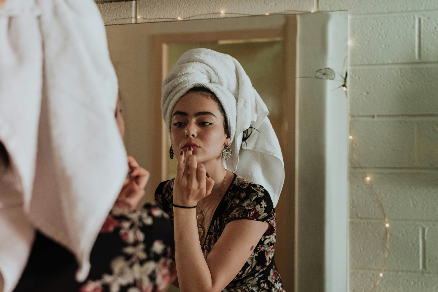 A woman wearing a black floral print top with a white towel on her head looks at her reflection in the mirror, using her ring finger to dab color onto her lips, with a white brick wall adorned with fairy lights visible around the mirror, highlighting the allure of high-quality skincare products for healthy skin.