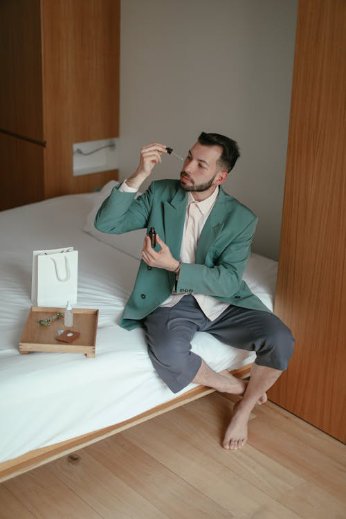 A man in a casual suit sitting on a bed, applying a skincare product using a dropper.