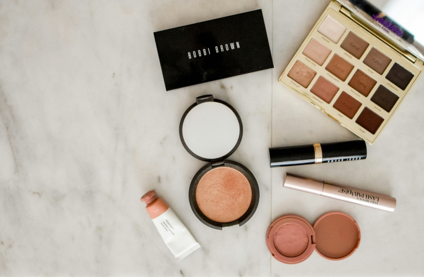  A makeup palette with neutral shades, a black Bobbi Brown box, an open blush case with a mirror, along with a lipstick, concealer pen, tube, and lip color case, arranged on a white and grey marble surface, showing that people tend to buy quality skincare products when it comes to makeup.