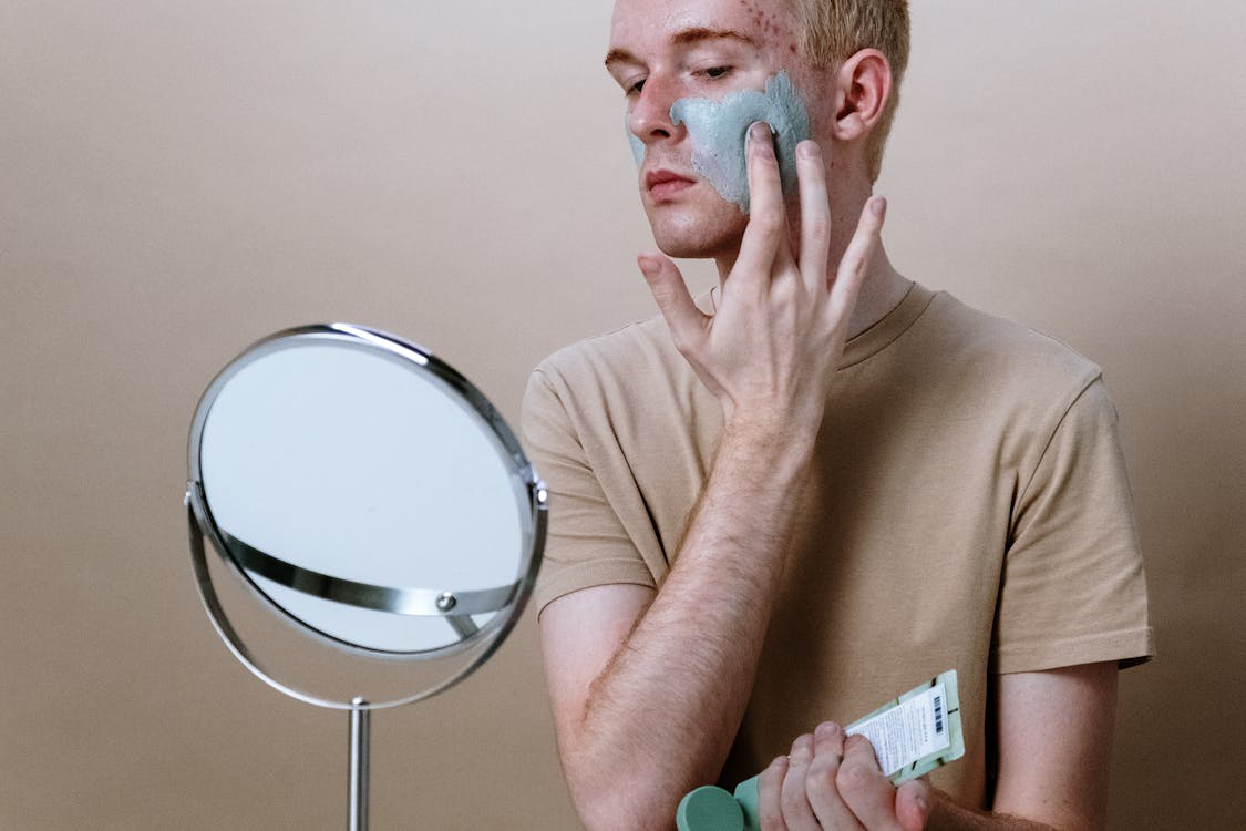 A man applying a facial mask while looking into a mirror, engaging in a modern skincare routine that challenges traditional masculinity.